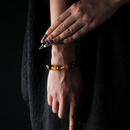 A close-up of a person’s hand against a black textured dress, wearing a beaded bracelet featuring glossy golden tiger’s eye, reddish and yellow-brown stones, accented with a transparent citrine bead. The bracelet creates a warm, earthy contrast under soft lighting, highlighting the natural luster of each polished bead.