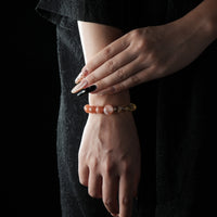 The image features a fashion model wearing the bracelet on their left wrist, with hands gently posed. The bracelet’s orange and golden tones complement the model’s dark, elegant outfit. The lighting subtly catches the gemstone reflections, giving the accessory a warm, radiant appearance.
