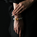 A close-up of a woman's hands elegantly resting on her wrist, showcasing a natural crystal bracelet composed of obsidian, strawberry quartz, grey moonstone, and yellow tiger’s eye beads. The woman wears a black textured dress, and the background is dark, highlighting the subtle glow and contrast of the stones.