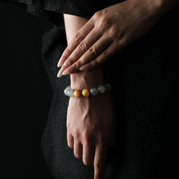 A close-up of a woman's hands elegantly resting on her wrist, showcasing a natural crystal bracelet composed of obsidian, strawberry quartz, grey moonstone, and yellow tiger’s eye beads. The woman wears a black textured dress, and the background is dark, highlighting the subtle glow and contrast of the stones.