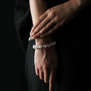 A refined close-up of a woman's wrist and hand, adorned with a natural crystal bracelet composed of strawberry quartz, clear quartz, citrine, and grey moonstone beads. Her nails are long and polished, and she is dressed in an elegant black textured outfit. The dark background and soft lighting emphasize the gentle hues and natural shine of the stones.