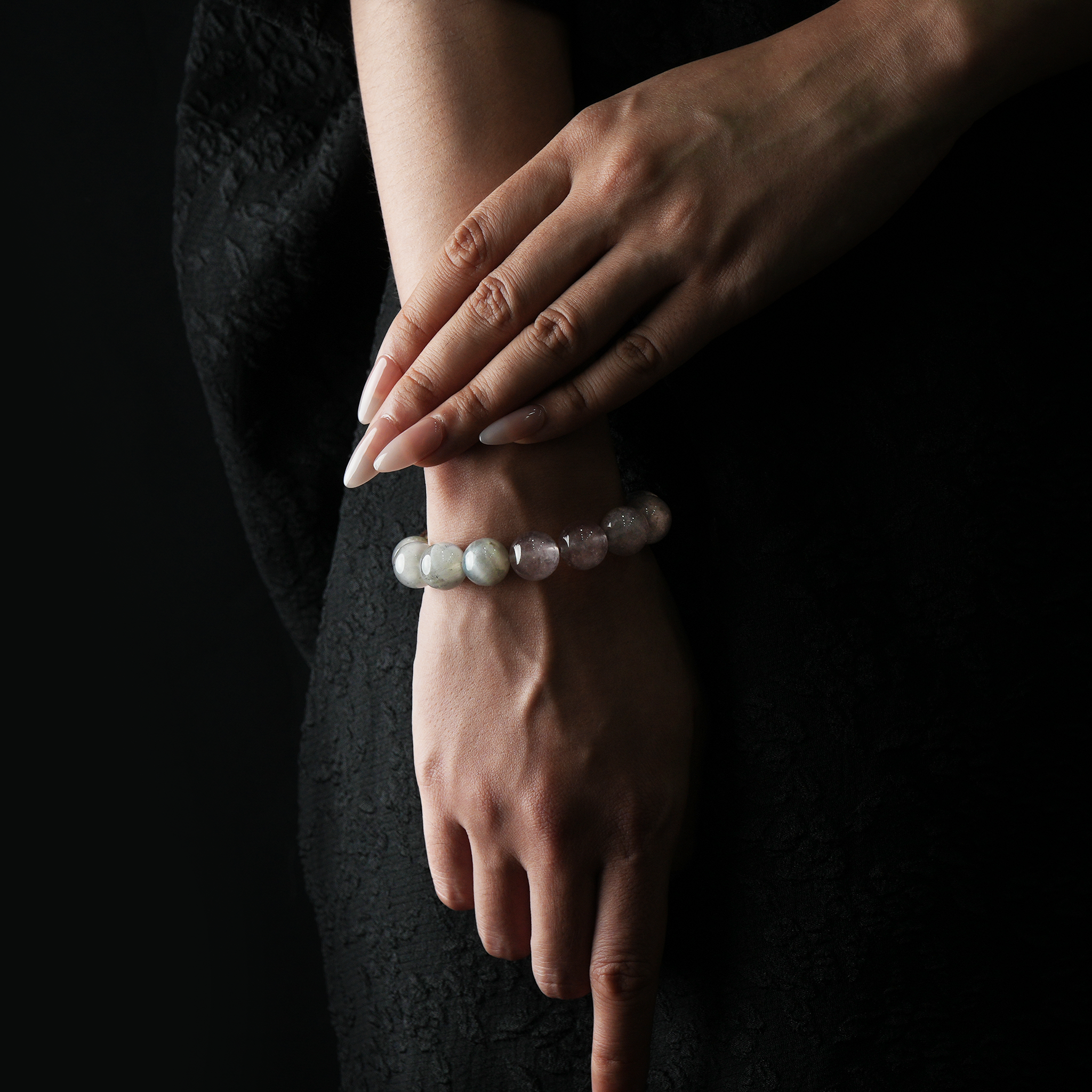 A refined close-up of a woman's wrist and hand, adorned with a natural crystal bracelet composed of strawberry quartz, clear quartz, citrine, and grey moonstone beads. Her nails are long and polished, and she is dressed in an elegant black textured outfit. The dark background and soft lighting emphasize the gentle hues and natural shine of the stones.