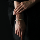 A close-up of a woman's wrist adorned with a slim, multicolored crystal bracelet featuring silver sheen obsidian, grey moonstone, strawberry quartz, and citrine beads. The beads form a soft gradient from dark to bright tones. Her hand is elegantly posed against a black textured dress, and the overall lighting is subdued, emphasizing the bracelet’s gentle luster and feminine refinement.