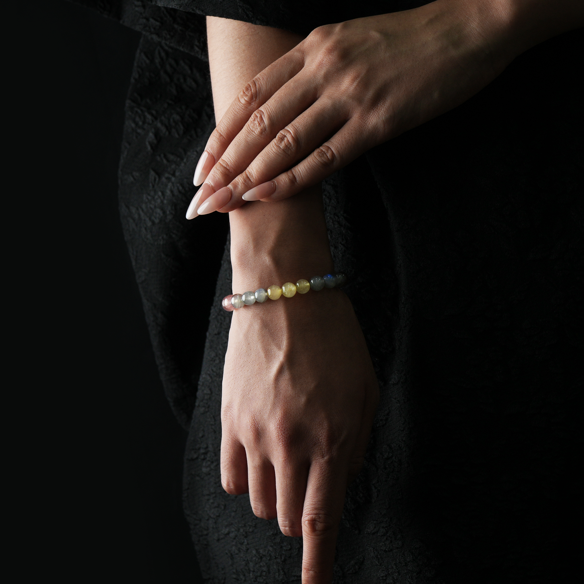 A close-up of a woman's wrist adorned with a slim, multicolored crystal bracelet featuring silver sheen obsidian, grey moonstone, strawberry quartz, and citrine beads. The beads form a soft gradient from dark to bright tones. Her hand is elegantly posed against a black textured dress, and the overall lighting is subdued, emphasizing the bracelet’s gentle luster and feminine refinement.