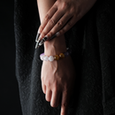 A close-up of a hand wearing a beaded bracelet made of Amethyst, Rose Quartz, and Yellow Tiger Eye. The soft lavender and blush tones are accented by warm golden-brown beads, creating a gentle yet eye-catching color contrast against a black textured outfit.