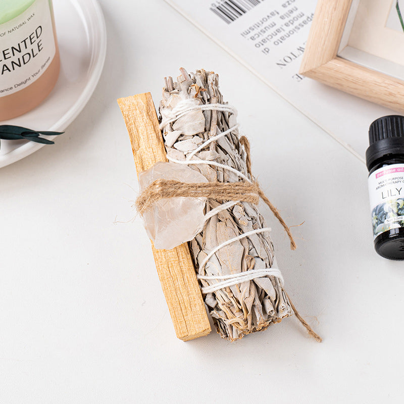 A sage and palo santo bundle tied with amethyst crystal next to a scented candle, on a soft tabletop background.