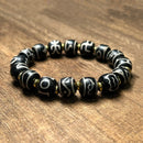 A Tibetan-style mantra bead bracelet displayed in a slightly angled view on a wood background. The bracelet features multiple black beads adorned with distinct white Tibetan symbols, separated by small gold spacers, representing a mix of spiritual symbolism and rustic elegance.