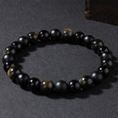 Bracelet laid flat on dark surface, composed of matte black agate and reflective gold sheen obsidian beads, offering strength and emotional shielding.
