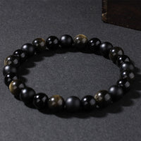 Bracelet laid flat on dark surface, composed of matte black agate and reflective gold sheen obsidian beads, offering strength and emotional shielding.