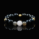 A beaded bracelet made of glossy black obsidian, shimmering gold hematite, and a central soft white moonstone, arranged in a symmetrical design and displayed on a reflective black surface for a sleek, elegant look.