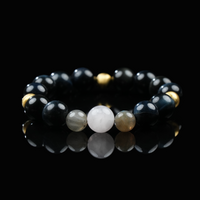 A beaded bracelet made of glossy black obsidian, shimmering gold hematite, and a central soft white moonstone, arranged in a symmetrical design and displayed on a reflective black surface for a sleek, elegant look.