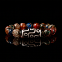 A striking black and white 9-eye Dzi bead sits at the center of a handmade bracelet, surrounded by glossy multicolored agate beads in shades of red, blue, bronze, and emerald. The bracelet is arranged in a circle against a black reflective background, emphasizing its vibrant yet balanced energy.