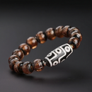 A spiritual bracelet centered around a cylindrical 9-eye Dzi bead in black and white, complemented by round, translucent amber-colored beads with metallic luster. The contrast between the mystical core bead and the fiery brown tones makes this a standout accessory.
