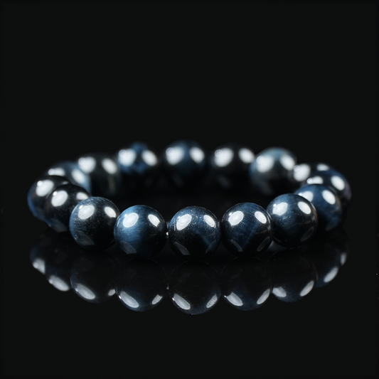 Close-up of a polished blue tiger eye bracelet placed on a black reflective surface, showcasing its deep navy sheen and layered chatoyancy under soft lighting.