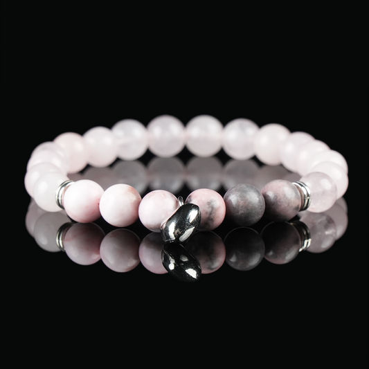 A close-up of a rose quartz and rhodonite beaded bracelet with a silver heart charm, placed on a reflective black surface.