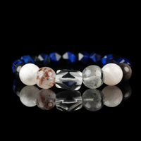 A gemstone bracelet featuring a central carved clear quartz bead, flanked by mica red rutilated quartz, snow phantom, silver obsidian, and faceted lapis lazuli beads. The arrangement of natural stones showcases a harmonious blend of translucent, earthy, and deep blue hues, set against a reflective black surface.