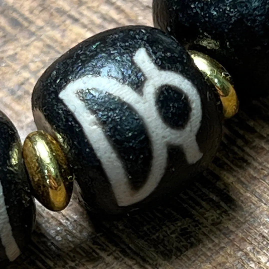 Detailed macro shot of a black Dzi bead showcasing a white Tibetan-style script, paired with a metallic gold spacer bead. The bead’s matte texture and precise hand-etched design highlight the craftsmanship and spiritual aesthetic of traditional Tibetan jewelry.