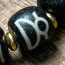 Detailed macro shot of a black Dzi bead showcasing a white Tibetan-style script, paired with a metallic gold spacer bead. The bead’s matte texture and precise hand-etched design highlight the craftsmanship and spiritual aesthetic of traditional Tibetan jewelry.