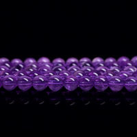 This image features a close-up view of multiple strands of polished deep purple crystal round beads, likely amethyst. The beads are arranged in neat rows on a glossy black surface, with a reflective finish that enhances their vibrant color and smooth texture.