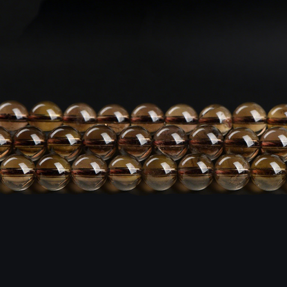 A close-up image showing multiple strands of polished smoky quartz round beads laid out horizontally. The beads display a translucent brown hue with a smooth, glossy surface and visible internal textures.