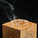 Close-up of gemstone bracelet on bamboo lid, smoke curling in soft light above it.