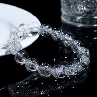 A Himalayan quartz bracelet displayed near a clear cup on a glossy black surface, reflecting vibrant light sparkles.