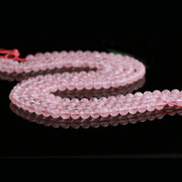 Strands of light pink rose quartz beads are arranged in an elegant curve across a glossy black background. The soft pink thread running through the beads is slightly visible, complementing their natural hue.