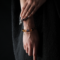 A person wears a handcrafted bracelet made of flame-inspired energy beads in amber and black hues, accented with gold-tone metal components. The design features a central purple bead and a dangling charm, blending elegance with spiritual symbolism.