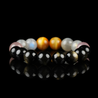 A reflective black surface displays a crystal bracelet arranged in a circle. The bracelet features dark black Obsidian beads, blush pink Strawberry Quartz, pearly Grey Moonstone, and radiant golden Tiger’s Eye stones, creating a balanced and earthy color palette with a spiritual vibe.