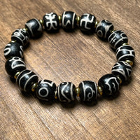 A circular arrangement of a handcrafted Tibetan Dzi bead bracelet resting on a wooden tabletop. The black beads, each inscribed with unique white script or symbols, are interspaced with golden spacers. The image emphasizes the symmetry, spiritual meaning, and earthy tone of the design.