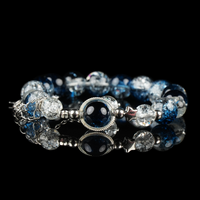 A celestial-inspired bracelet featuring galaxy energy beads in deep blue and crackled clear tones, accented with silver star spacers and a central orb framed in silver. The reflective black surface enhances its cosmic and mystical aesthetic.