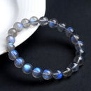 Gray moonstone bracelet casually resting near a white dish, with multiple beads showing distinct flashes of blue under soft lighting.