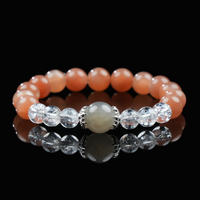 A beaded bracelet made of shimmering gray moonstone at the center, flanked by clear quartz and warm orange moonstone beads, displayed on a black reflective surface, held gracefully by hand, and elegantly worn on the wrist.