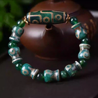 This vibrant green Dzi bead bracelet is draped around a traditional brown teapot, highlighting the gold-ringed circular motifs on each bead. The main elongated Dzi bead carries a rich golden 9-eye design, signifying wisdom and longevity, contrasting beautifully with the deep green tones of the surrounding stones.
