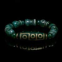 A handcrafted green agate Dzi bead bracelet placed on a glossy black surface. The central bead features a traditional 9-eye design in gold, symbolizing protection and spiritual insight, and is surrounded by symmetrical round and barrel-shaped accent beads, creating a sense of cultural depth and elegance.