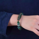 Captured on a wrist, the bracelet's elegance is accentuated by the contrast with a deep navy-blue sleeve. The large central Dzi bead, inscribed with a golden 9-eye pattern, sits prominently atop the wrist, while alternating green and turquoise-toned beads enhance its rich, spiritual character.