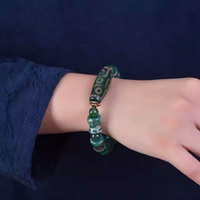 Captured on a wrist, the bracelet's elegance is accentuated by the contrast with a deep navy-blue sleeve. The large central Dzi bead, inscribed with a golden 9-eye pattern, sits prominently atop the wrist, while alternating green and turquoise-toned beads enhance its rich, spiritual character.