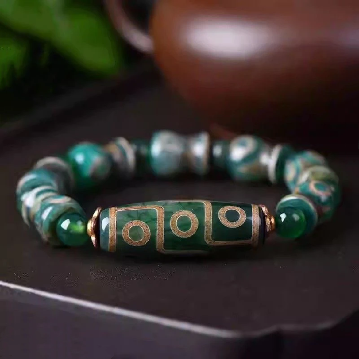 The bracelet lies elegantly on a dark surface, with its central elongated Dzi bead showcasing bold circular and linear golden patterns against a rich green base. The supporting beads alternate between polished green agate rounds and uniquely shaped Tibetan barrel beads, evoking an aura of tradition and spiritual symbolism.