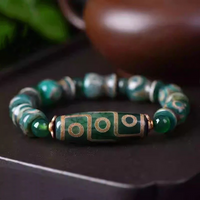 The bracelet lies elegantly on a dark surface, with its central elongated Dzi bead showcasing bold circular and linear golden patterns against a rich green base. The supporting beads alternate between polished green agate rounds and uniquely shaped Tibetan barrel beads, evoking an aura of tradition and spiritual symbolism.