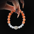 A close-up image showing a hand with long manicured nails holding a round beaded bracelet composed of Gray Moonstone, Orange Moonstone, and Clear Quartz beads against a black background.