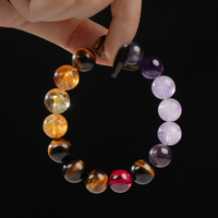 A close-up image of a hand delicately holding a circular beaded bracelet composed of vibrant natural crystals including purple amethyst, golden citrine, deep red garnet, and golden-brown tiger’s eye. The stones are highly polished and arranged in a harmonious gradient, presented against a sleek black backdrop.