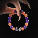 A close-up of a hand with long, manicured nails holding a beaded bracelet made of alternating Amethyst and Orange Moonstone stones, featuring a detailed silver butterfly charm at the center.