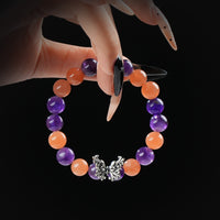 A close-up of a hand with long, manicured nails holding a beaded bracelet made of alternating Amethyst and Orange Moonstone stones, featuring a detailed silver butterfly charm at the center.