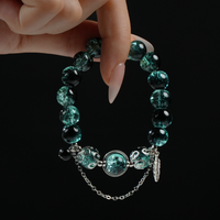 A hand displaying a handmade forest-themed bracelet with cracked green beads, silver feather charm, and grounding aesthetic, blending elegance and earthy strength.