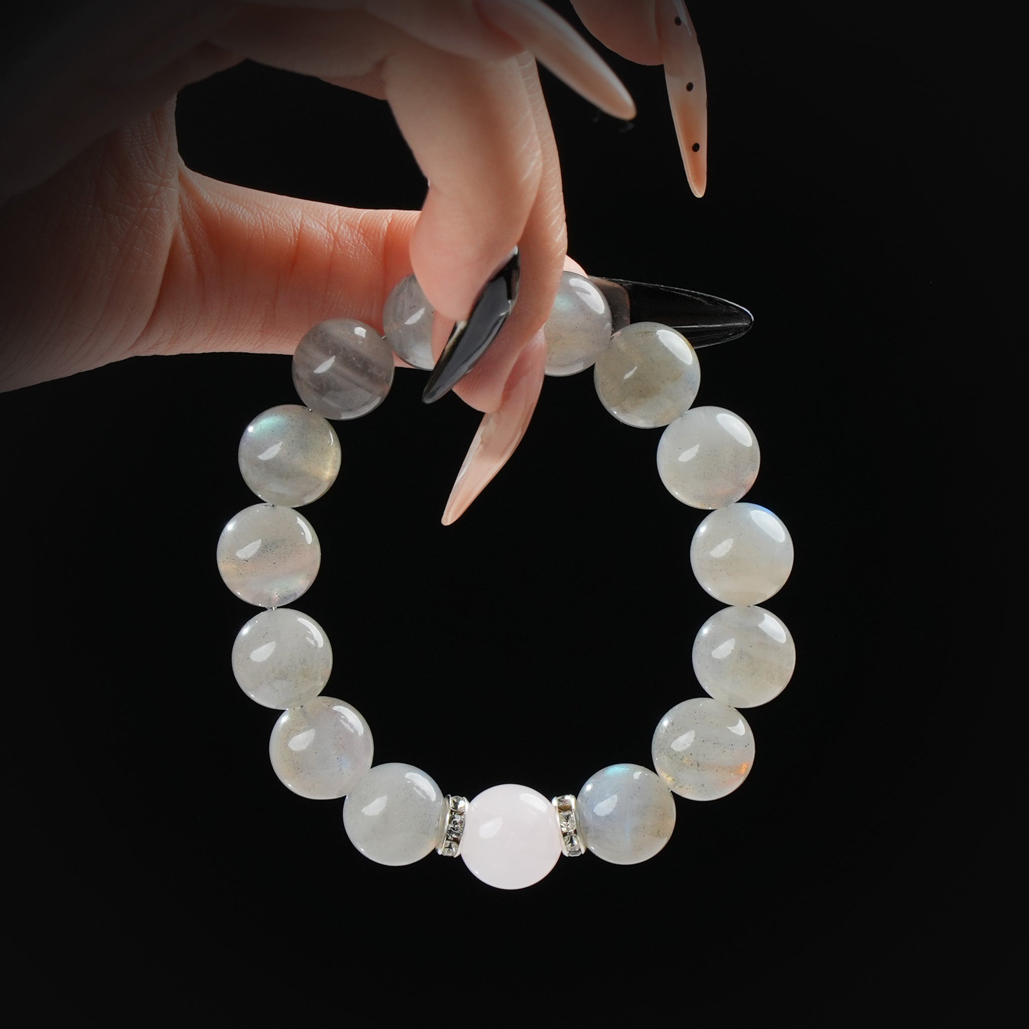 Delicate fingers holding a handmade bracelet of rose quartz and gray moonstone beads, emphasizing the soft pink centerpiece against dark