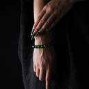 A close-up shot of a woman's hand and wrist wearing a tricolor crystal bracelet made of green agate, amethyst, and tiger eye beads. The deep green, rich purple, and golden-brown beads contrast elegantly against a black textured dress background, giving the bracelet a natural yet refined visual appeal.
