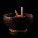 A small coconut bowl on a rope base, containing herbs and a gemstone bracelet under soft lighting.
