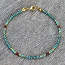 A close-up of a thin turquoise anklet with red, gold, and blue accents on a textured wooden background.