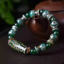 The bracelet is shown from a dynamic side angle, coiled gently over a teapot, enhancing the bracelet’s three-dimensional texture. The alternating bead shapes and the intricate gold inlay on the Dzi bead reflect traditional Tibetan artistry and spiritual meaning associated with abundance and protection.
