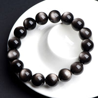 A silver sheen obsidian bracelet coiled on white porcelain, reflecting soft light from its surface.